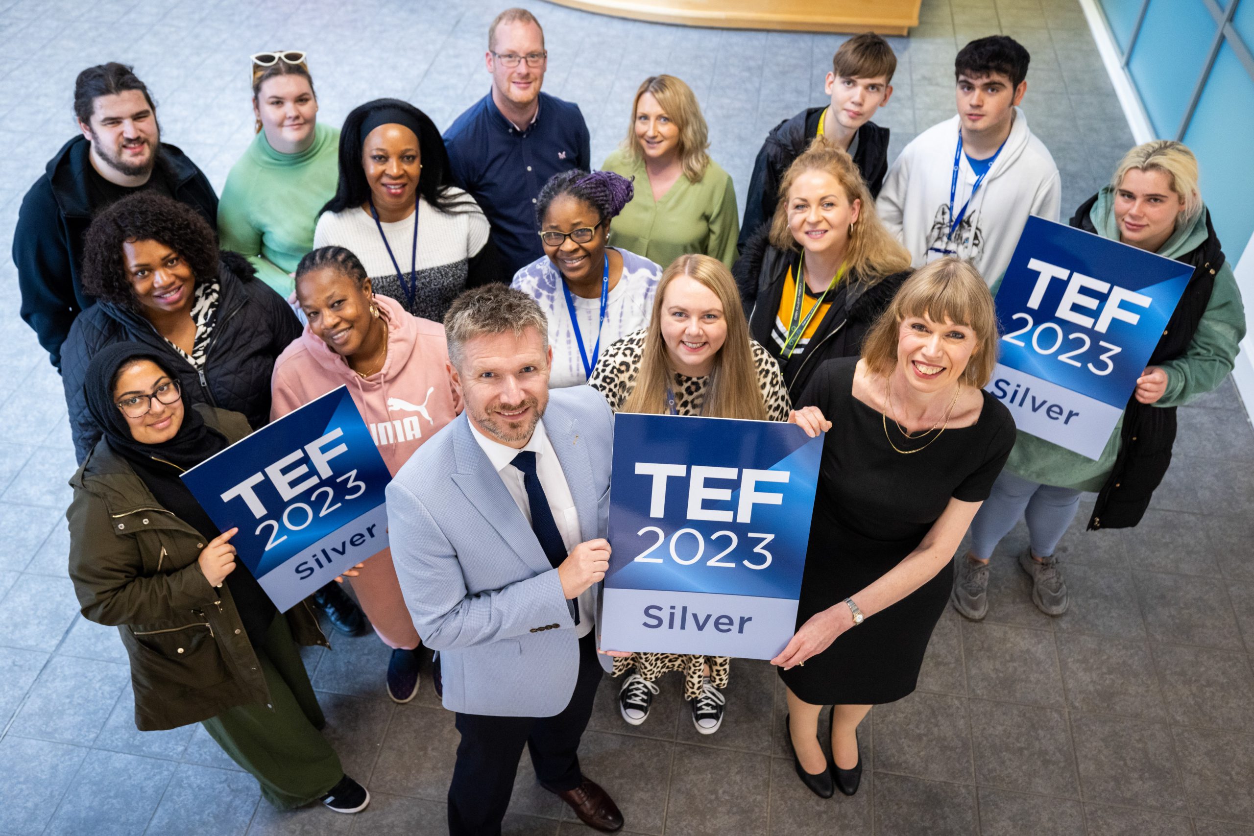 Students and staff smile at the camera, holding TEF 2023 Silver posters.