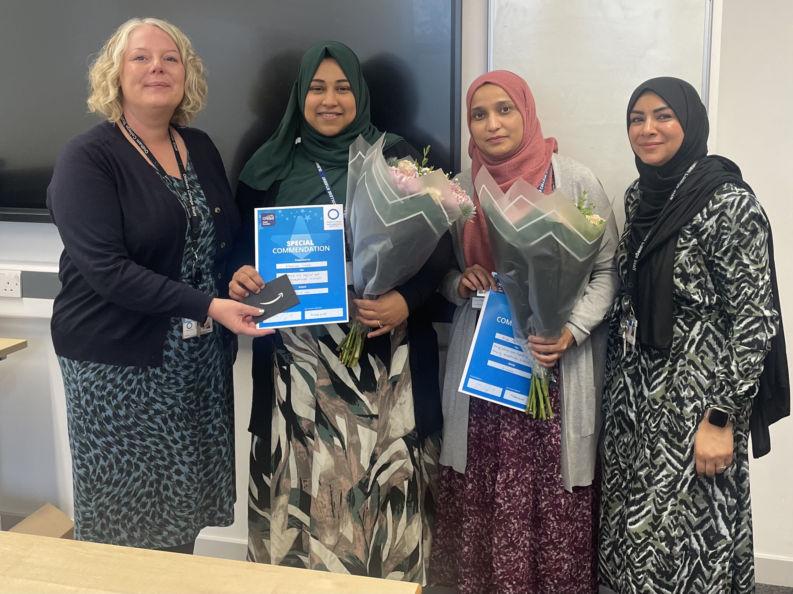 Four people stand in a row; the two people in the middle each hold a bouquet of flowers and blue certificate.