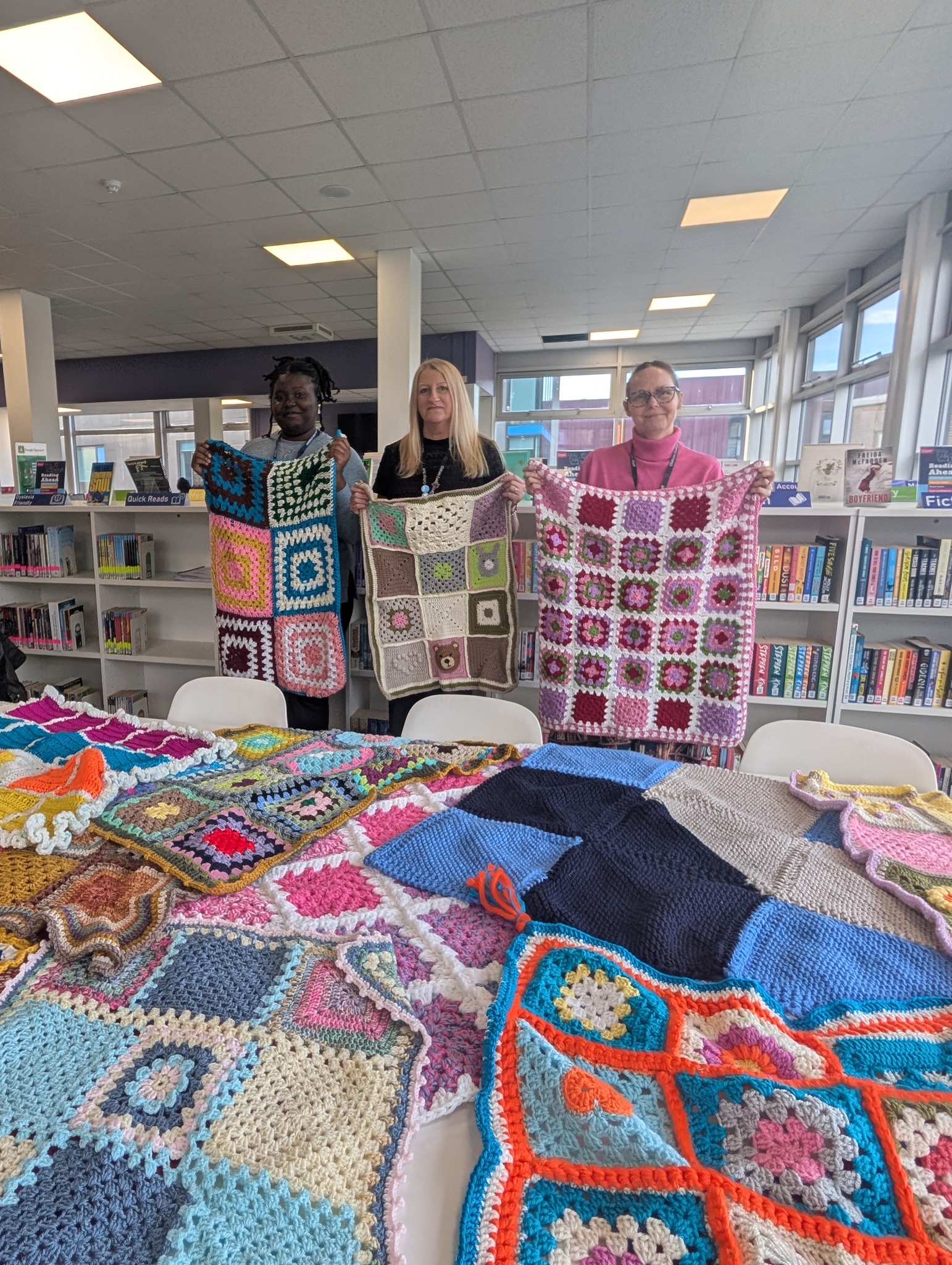 Three people stand behind a table laid with crocheted blankets in a library. They each hold a blanket