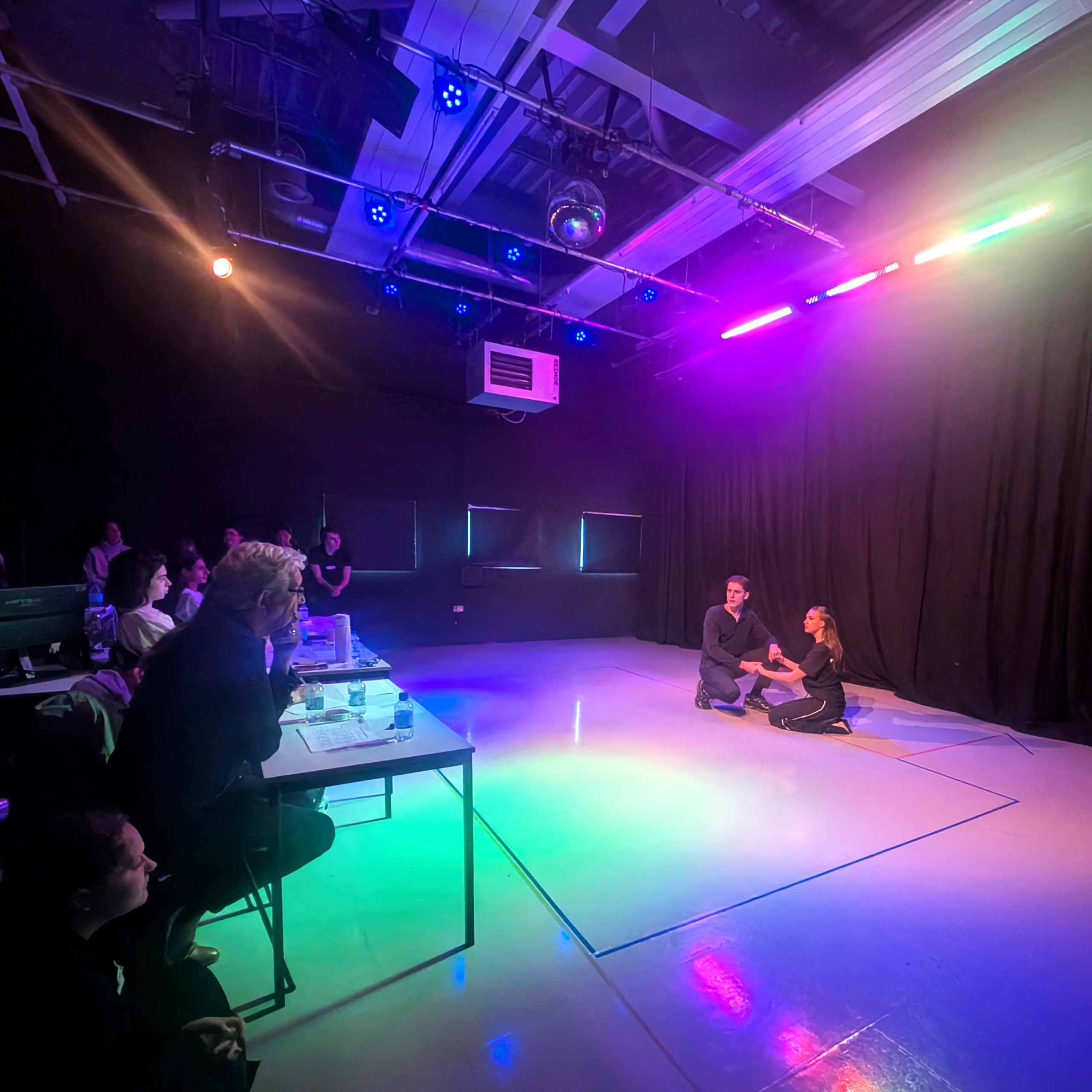 Two people sit on the floor in a room lit with coloured lights, delivering a performance, in front of an audience sitting on chairs behind tables