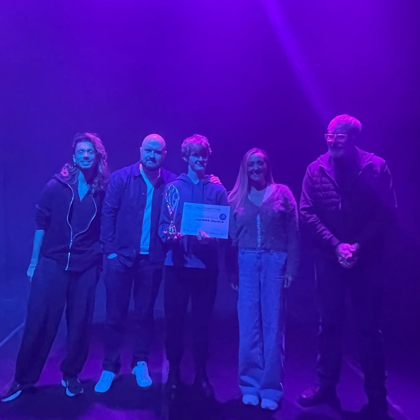 A group of five people stand in a room lit by purple lights. The person in the middle holds a certificate.