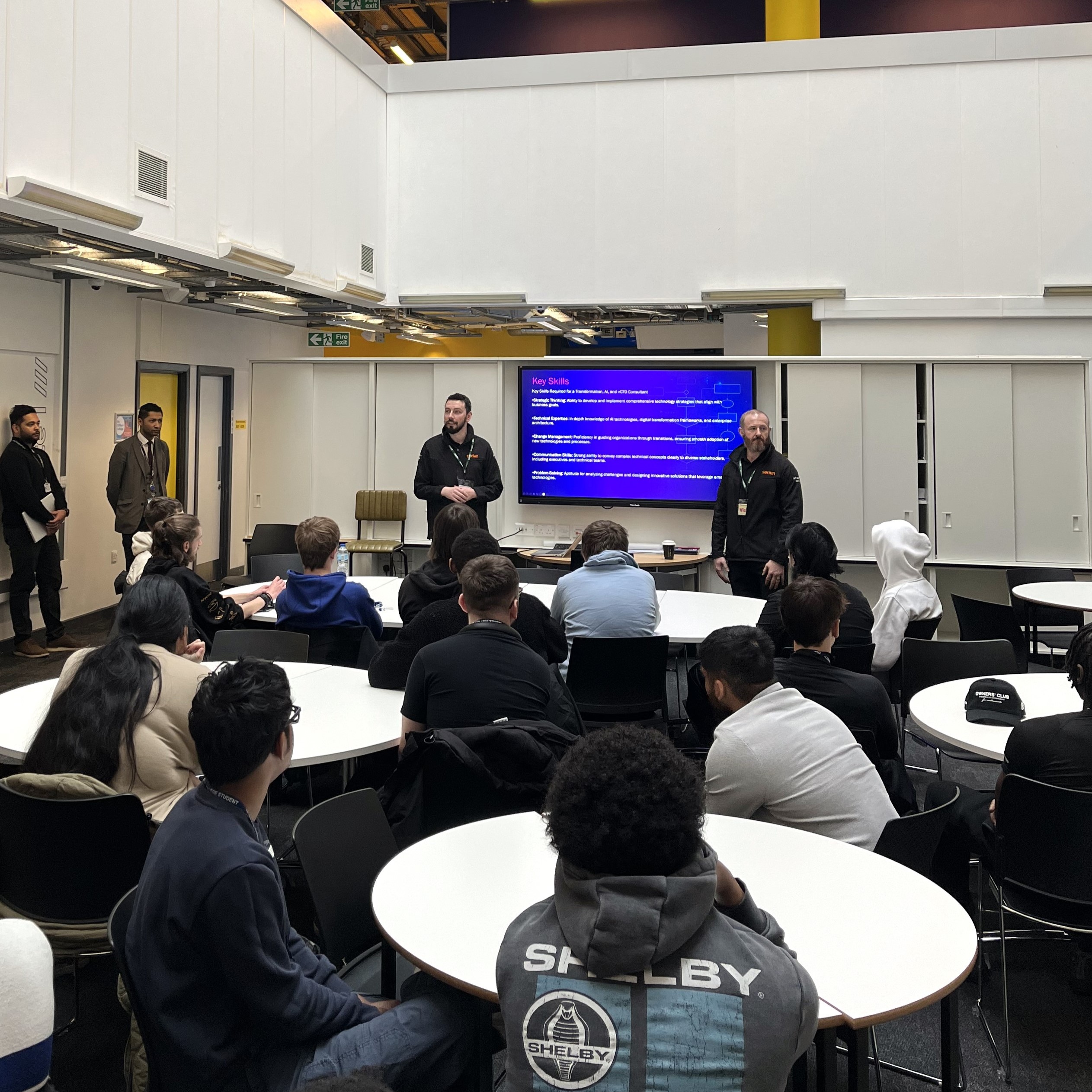 Two people give a talk to a classroom of people sitting at tables